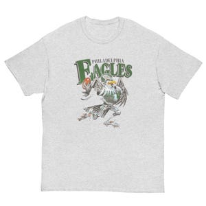Peut inclure: Un t-shirt gris clair à manches courtes avec un motif d'aigle portant un uniforme de football américain et tenant un ballon. Au-dessus de l'aigle, le texte Philadelphia Eagles est inscrit en lettres vert foncé avec un contour blanc.
