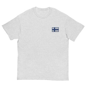 Peut inclure: Un t-shirt gris clair avec un petit drapeau finlandais brodé sur la poitrine gauche. Le drapeau présente une croix nordique bleue sur fond blanc. Le t-shirt a un col rond et des manches courtes.