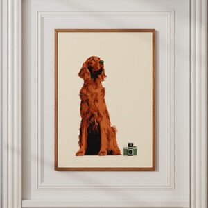 May include: A framed wall art print featuring a solid cream background with a detailed illustration of a long-haired, reddish-brown Irish Setter dog sitting upright. Beside the dog sits a small, vintage-style green twin-lens reflex camera.
