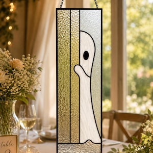 May include: A rectangular stained glass panel featuring a white ghost figure peeking from behind a vertical gold textured glass strip. The piece is framed in black metal and hangs from a black chain, set against a clear, textured glass background.