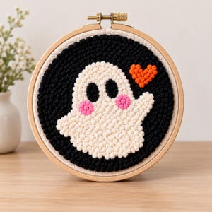 May include: A circular wooden embroidery hoop containing a punch needle art piece. The design features a white ghost with black eyes and pink cheeks against a solid black background, accompanied by a small orange heart shape to the right.