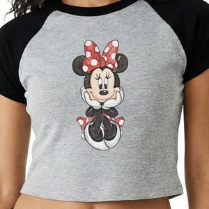 May include: A gray crop top with black sleeves featuring a Minnie Mouse graphic. The cartoon character is depicted with a red and white polka dot bow, black ears, and a red and white polka dot skirt. The shirt is worn by a person.
