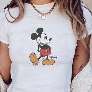 May include: White t-shirt featuring a graphic of Mickey Mouse in a walking pose. The cartoon character is in black, red, and yellow. The Disney logo is printed below the character.