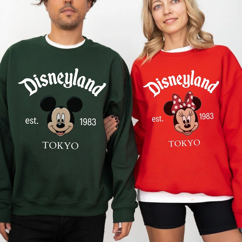 Pu&ograve; includere: Due felpe, una verde scuro e una rossa, con il logo Disneyland e Topolino e Minnie. Il testo include "est. 1983" e "TOKYO". La felpa verde ha Topolino, e quella rossa ha Minnie.