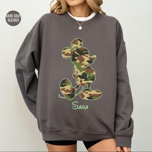 May include: Dark gray heather sweatshirt featuring a camouflage Mickey Mouse design. The iconic character is filled with green, brown, and tan camouflage pattern. The name "Sara" is written in green script below the character. The text "Dark Gray Heather" is in the upper left corner.