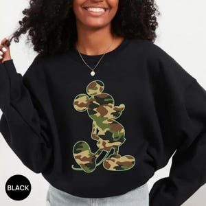 May include: Black crewneck sweatshirt featuring a camouflage Mickey Mouse graphic. The Mickey Mouse design incorporates shades of green, brown, and tan. The word "BLACK" is printed on the bottom left of the image.