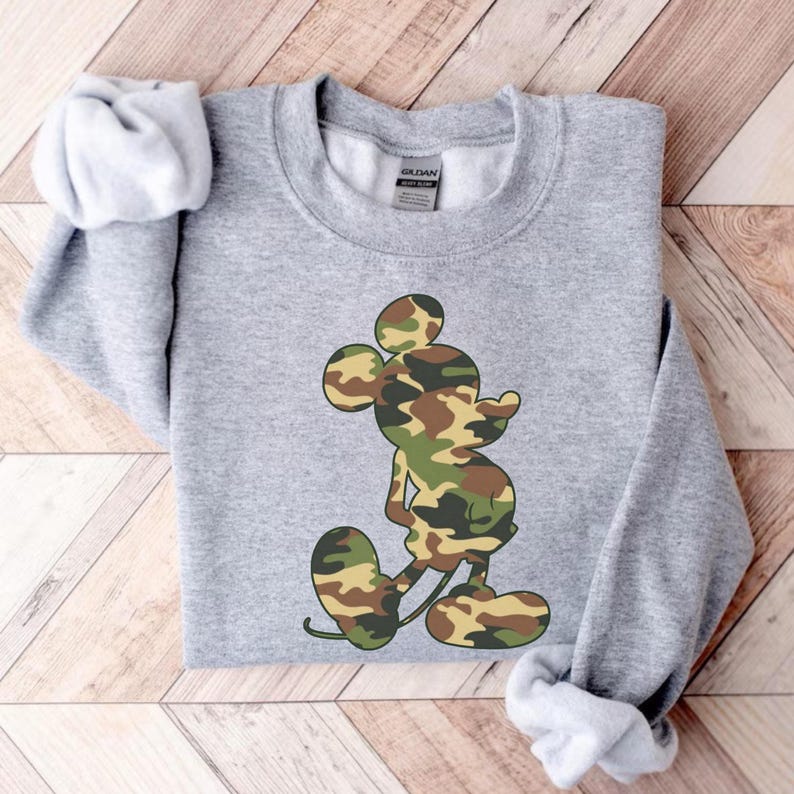 May include: A gray sweatshirt featuring a camouflage Mickey Mouse design. The graphic uses shades of green, brown, and tan. The sweatshirt has a crew neck and long sleeves with cuffed wrists. The fabric appears soft and comfortable.