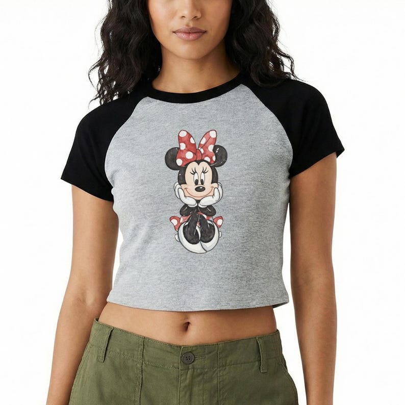 May include: A gray crop top with black sleeves featuring a Minnie Mouse graphic. The cartoon character is depicted with a red and white polka dot bow, black ears, and a red and white polka dot skirt. The shirt is worn by a person.