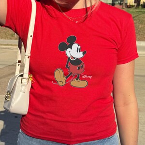 May include: A red t-shirt featuring a graphic of Mickey Mouse in a walking pose. The cartoon character is depicted with black ears, a white face, red shorts, and brown shoes. The Disney logo is printed below the character.