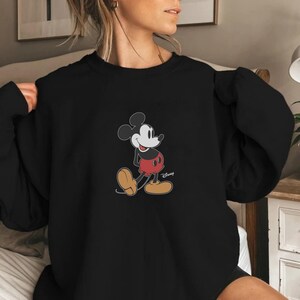 Pu&ograve; includere: Felpa nera con grafica di Topolino. Il personaggio dei cartoni animati &egrave; raffigurato nella sua posa classica, con pantaloncini rossi, scarpe gialle e guanti bianchi. Il logo Disney &egrave; visibile in basso a destra.
