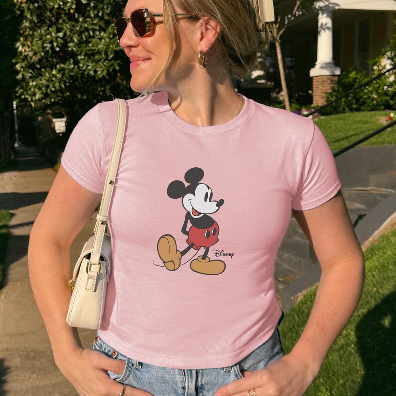 May include: A light pink t-shirt featuring a graphic of Mickey Mouse in his classic pose. The cartoon character is depicted in black, red, and yellow. The word "Disney" is printed below the character.