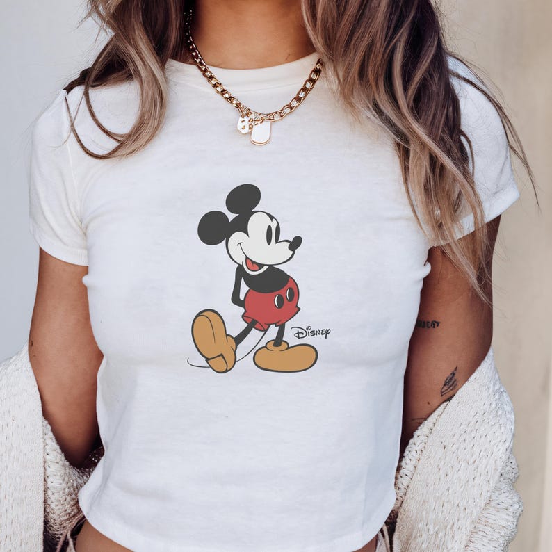 May include: White t-shirt featuring a graphic of Mickey Mouse in a walking pose. The cartoon character is in black, red, and yellow. The Disney logo is printed below the character.