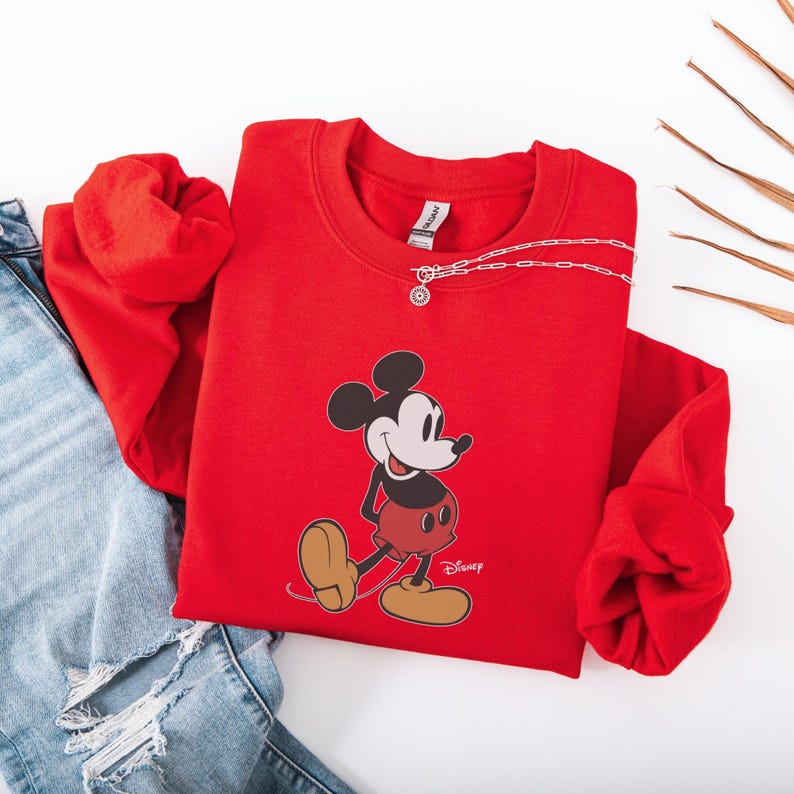Pu&ograve; includere: Una felpa rossa con una grafica di Topolino. La felpa ha girocollo e maniche lunghe. Una collana d'argento con un ciondolo &egrave; drappeggiata sul davanti. Il logo Disney &egrave; stampato in basso a destra.