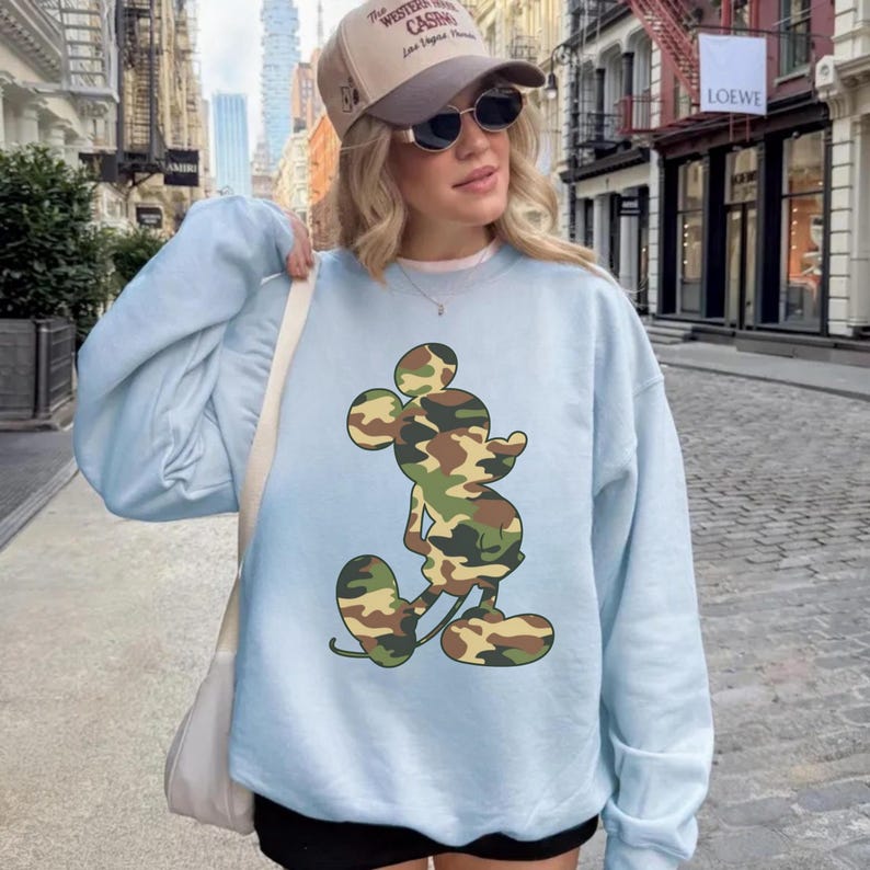 May include: Light blue sweatshirt featuring a camouflage Mickey Mouse graphic. The sweatshirt has long sleeves and a crew neck. The image also shows a baseball cap and sunglasses.