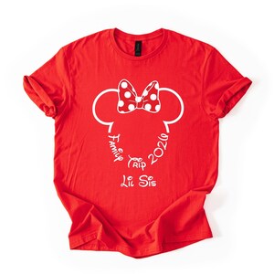 K&ouml;nnte beinhalten: Rotes T-Shirt mit einem wei&szlig;en Minnie-Mouse-Design. Das Design umfasst eine gepunktete Schleife, Mauseohren und die W&ouml;rter "Family Trip 2026" und "Lil Sis" in einer Schreibschrift.