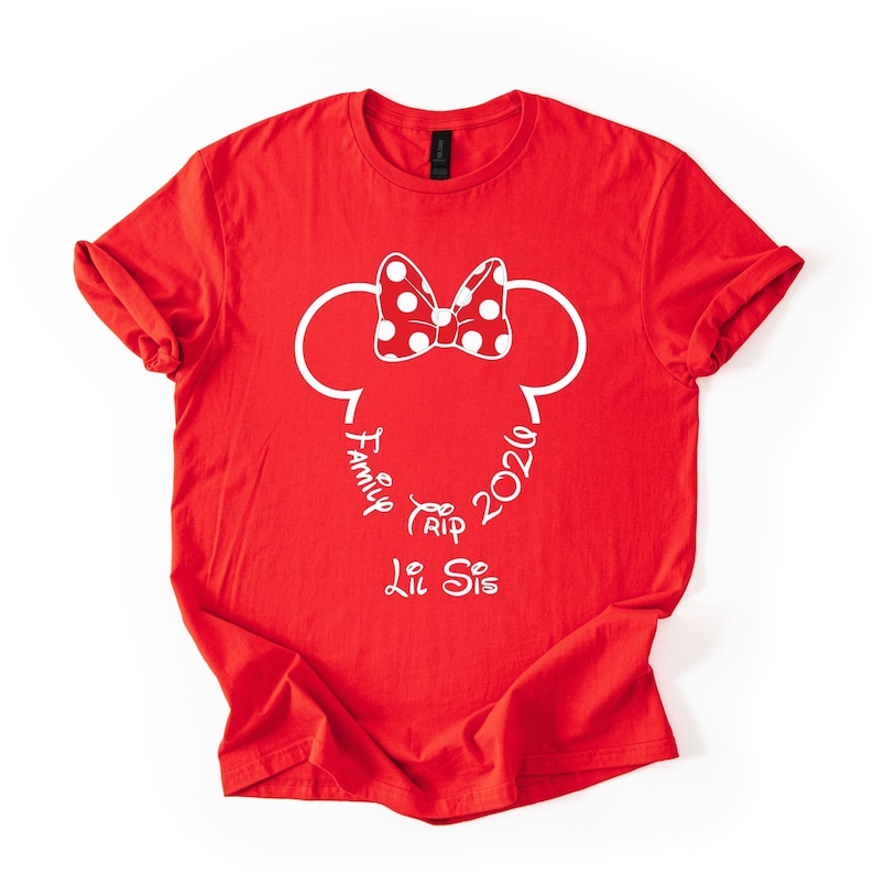 K&ouml;nnte beinhalten: Rotes T-Shirt mit einem wei&szlig;en Minnie-Mouse-Design. Das Design umfasst eine gepunktete Schleife, Mauseohren und die W&ouml;rter "Family Trip 2026" und "Lil Sis" in einer Schreibschrift.