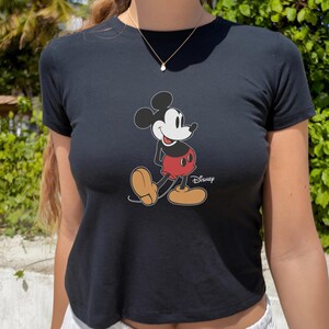 May include: Black crop top featuring a classic Mickey Mouse graphic. The cartoon character is depicted in his iconic pose, wearing red shorts and yellow shoes. The Disney logo is printed below the character.