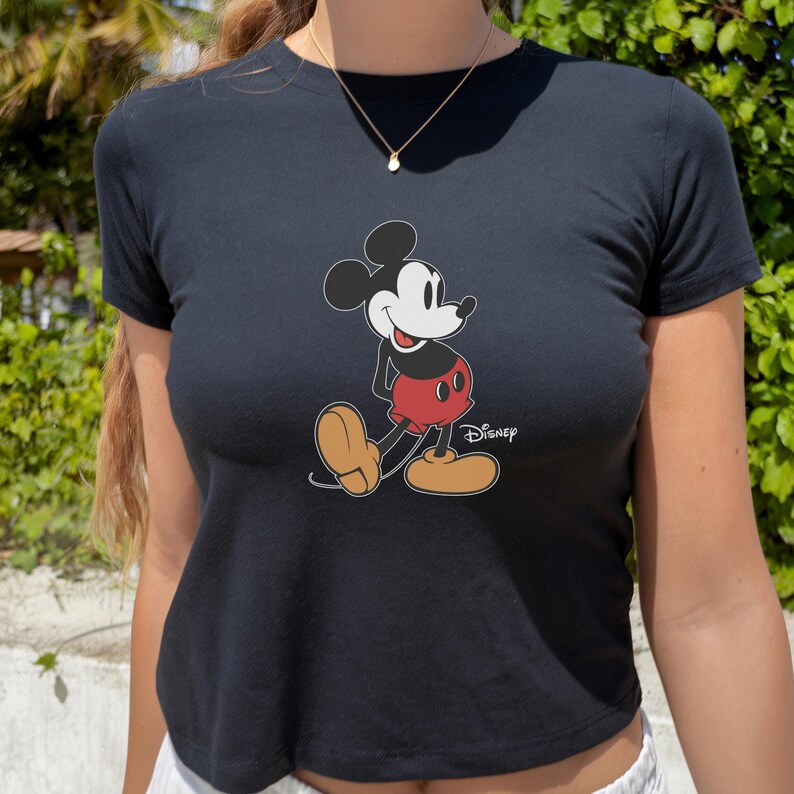May include: Black crop top featuring a classic Mickey Mouse graphic. The cartoon character is depicted in his iconic pose, wearing red shorts and yellow shoes. The Disney logo is printed below the character.