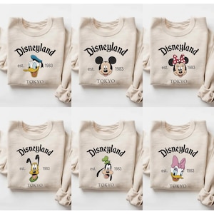 Pu&ograve; includere: Sei felpe beige con il logo Disneyland e vari personaggi Disney. Ogni felpa ha il testo "Disneyland est. 1983 TOKYO". I personaggi includono Paperino, Topolino, Minnie, Pluto, Pippo e Paperina.