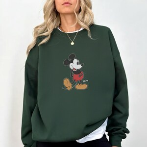 Pu&ograve; includere: Felpa verde scuro con grafica di Mickey Mouse. Il personaggio dei cartoni animati &egrave; raffigurato nella sua posa classica, con pantaloncini rossi e scarpe gialle. La felpa ha girocollo e maniche lunghe, con una maglietta bianca sotto.
