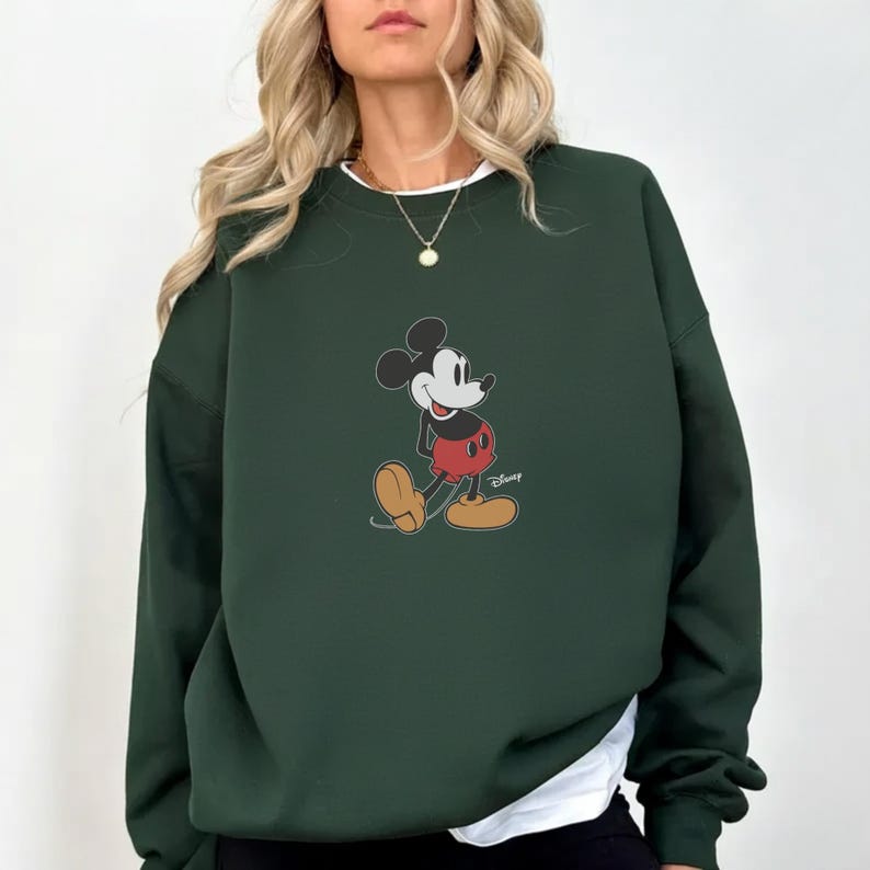 Pu&ograve; includere: Felpa verde scuro con grafica di Mickey Mouse. Il personaggio dei cartoni animati &egrave; raffigurato nella sua posa classica, con pantaloncini rossi e scarpe gialle. La felpa ha girocollo e maniche lunghe, con una maglietta bianca sotto.