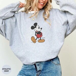 Pu&ograve; includere: Felpa grigio atletico con grafica di Mickey Mouse. Il personaggio dei cartoni animati indossa pantaloncini rossi, scarpe gialle e guanti neri. La felpa &egrave; abbinata a jeans blu. Il testo "ATHLETIC GRAY" &egrave; in basso a sinistra.