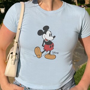 May include: Light blue t-shirt featuring a graphic of Mickey Mouse in his classic pose. The cartoon character is depicted in black, red, and tan colors. The Disney logo is printed on the bottom right.
