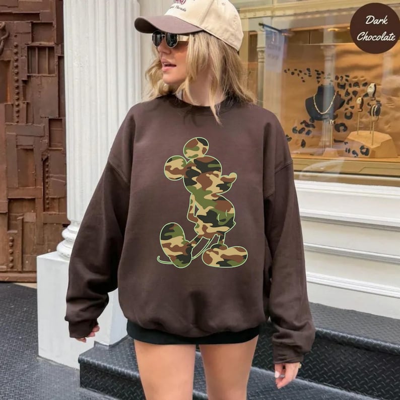 May include: Dark chocolate brown sweatshirt featuring a camouflage Mickey Mouse design. The sweatshirt has long sleeves and a crew neck. The text "Dark Chocolate" is in the upper right corner. The person in the image is wearing a baseball cap and sunglasses.