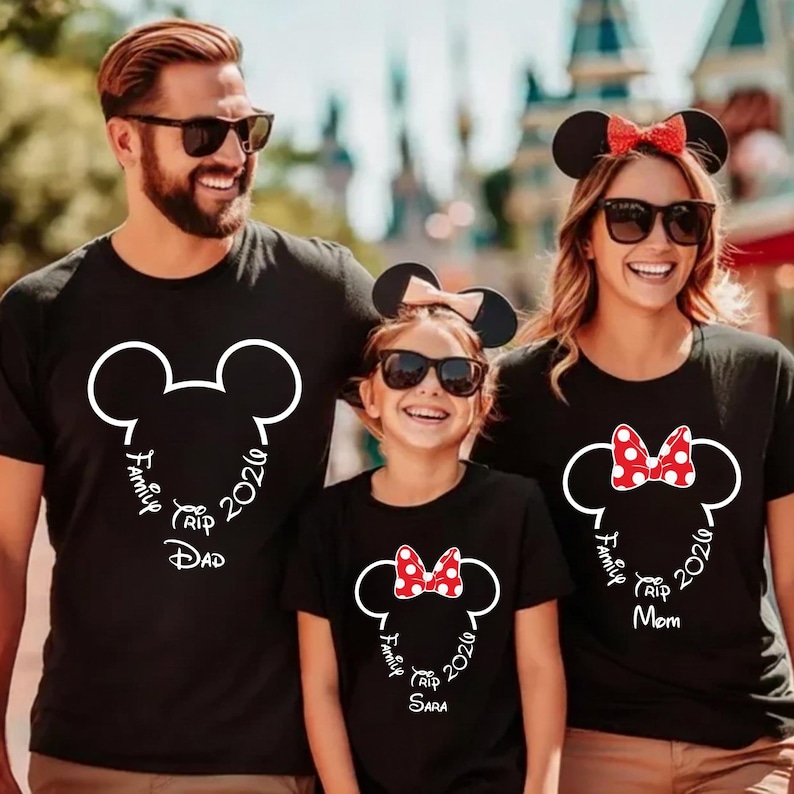 K&ouml;nnte beinhalten: Drei Personen tragen passende schwarze T-Shirts mit einem wei&szlig;en Mickey Mouse-Kopf-Design. Die Shirts tragen die Aufschrift "Family Trip 2026" und die W&ouml;rter "Dad", "Mom" und "Sara" unter dem Design. Die Familie tr&auml;gt Sonnenbrillen und M&auml;useohren.