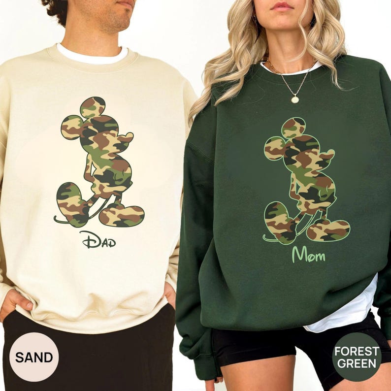 May include: Two sweatshirts, one sand-colored and one forest green, each featuring a camouflage Mickey Mouse design. The sand-colored sweatshirt has the word "Dad" printed below the character, while the forest green one says "Mom".