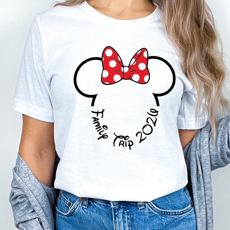 K&ouml;nnte beinhalten: Wei&szlig;es T-Shirt mit schwarzer Umrandung von Mickey Mouse Ohren und einer roten Schleife mit wei&szlig;en Punkten. Die Worte "Family Trip 2026" sind kreisf&ouml;rmig um die Ohren geschrieben.