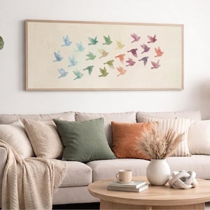 Puede incluir: Una obra de arte rectangular enmarcada que representa una bandada de pájaros de acuarela en tonos de azul, verde, amarillo, naranja y morado. La obra de arte se exhibe sobre un sofá beige con varias almohadas y una mesa de centro de madera.