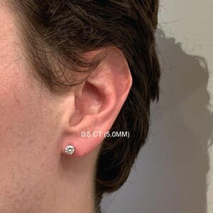 Op de afbeelding: Een close-up van een oor met een enkele ronde diamant stud oorbellen. De oorbel is 0,5 CT (5,0 mm) groot. De oorbel is gezet in een witte metalen zetting. De persoon heeft donkerbruin haar.