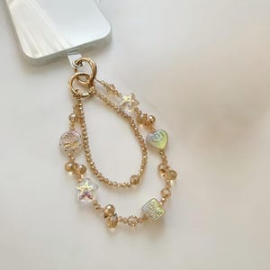 Könnte beinhalten: Ein goldfarbener Telefonanhänger mit einem doppelsträngigen Design. Der Anhänger verfügt über klare stern-, herz- und quadratförmige Perlen sowie kleinere bernsteinfarbene Perlen. Der Anhänger ist an einem goldfarbenen Ring befestigt.