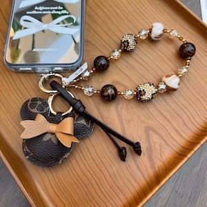 May include: A brown and tan leather Mickey Mouse-shaped keychain with a bow, attached to a beaded wrist strap. The strap features leopard print, clear, and brown beads. A gold-colored keyring is also present.