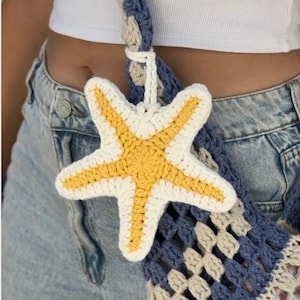 Puede incluir: Un charm de estrella de mar de ganchillo con centro amarillo y borde blanco cuelga de un bolso de ganchillo azul y crema. La imagen incluye el texto "Patrón de crochet + video instruction" y el identificador de Instagram "@margmer_crochet."