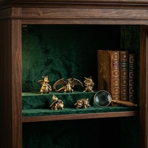 May include: A wooden cabinet interior with a green velvet backdrop displays several small, gold-colored dragon figurines. Also present are antique books and a magnifying glass, creating a fantasy theme.