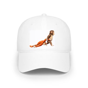 May include: White baseball cap featuring a cartoon illustration of two monkeys. One monkey is pulling the other by the arm. The illustration is in shades of brown and orange. The cap has a curved brim and an adjustable strap.