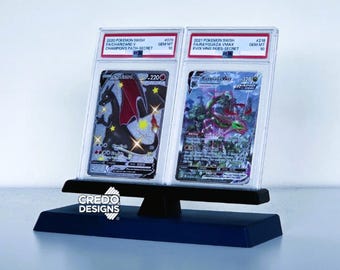 May include: A display stand holding two graded Pokémon trading cards. The cards are in protective cases, featuring Charizard and Rayquaza. The stand is black with a blue base, and the "CREDO DESIGNS" logo is visible.