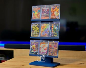 May include: A dark blue tiered display stand holding nine trading cards in clear protective cases. The stand features three horizontal shelves, each supporting three cards. A white logo with the text CREDO DESIGNS is visible on the base.