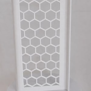 May include: A white, rectangular, upright panel featuring a repeating honeycomb or hexagonal cutout pattern. The structure has a smooth, solid border frame and a flat, vertical edge on the right side, presenting a clean, geometric design.