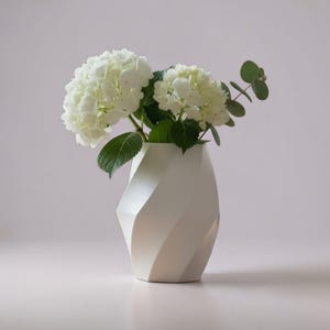 Op de afbeelding: Een witte, matte vaas met een geometrisch, spiraalvormig en gefacetteerd ontwerp. De vaas bevat twee grote, ronde, witte hortensiabloemen en een enkele tak met groene eucalyptusbladeren met klein, rond blad dat naar rechts uitsteekt.