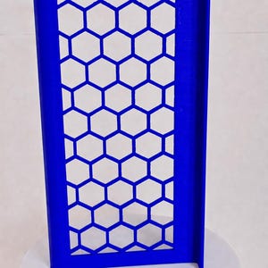 May include: A rectangular blue panel featuring a repeating honeycomb pattern of hexagonal cutouts. The panel has a solid frame and a perpendicular side flange, standing upright on a circular base. The structure is uniform in color and design.