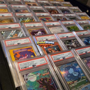 May include: A collection of graded trading cards in clear plastic cases arranged in rows. The cards feature colorful illustrations of various creatures and characters, with white labels displaying text, serial numbers, and PSA grading information.