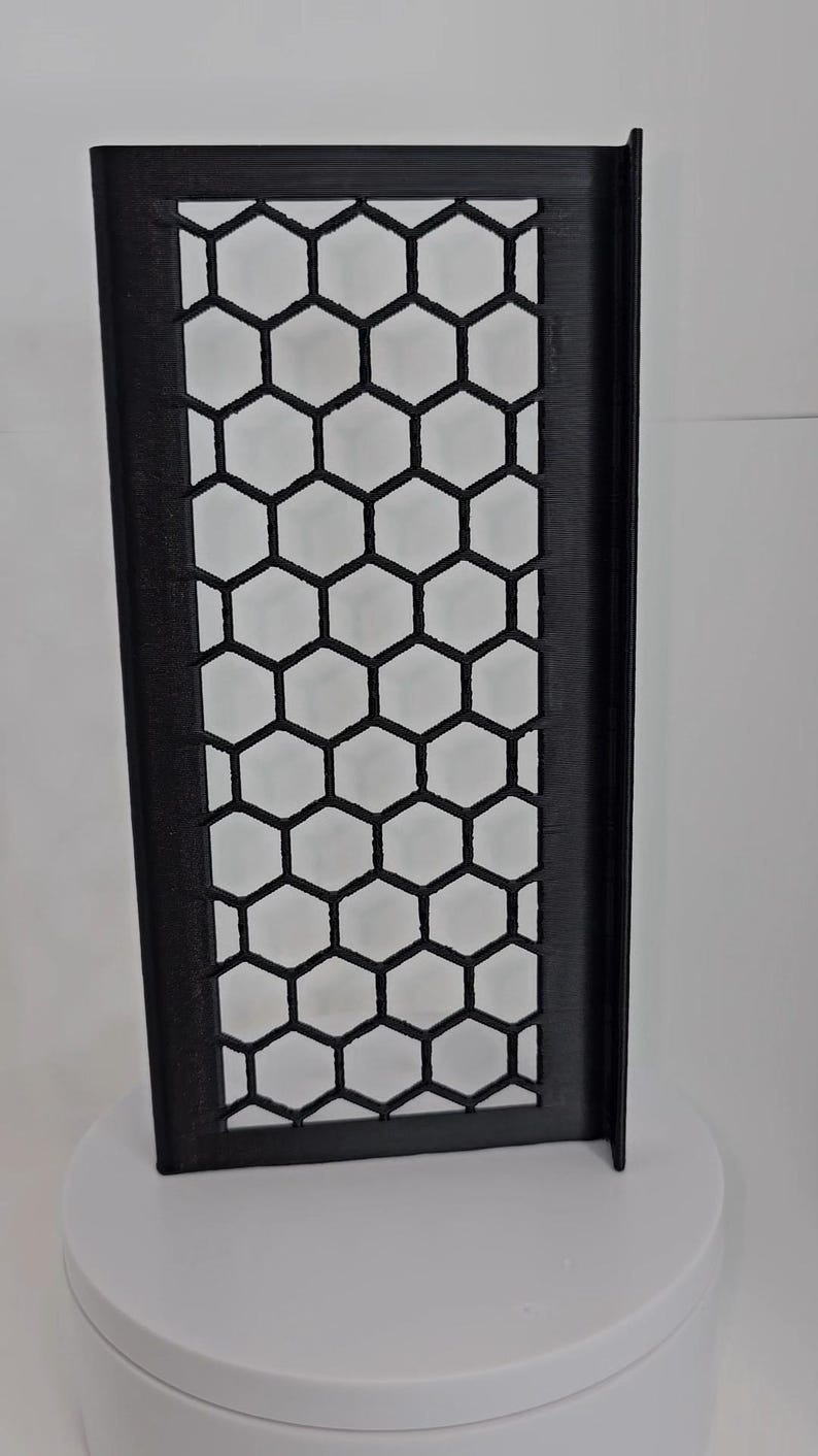 May include: A rectangular black panel featuring a repeating honeycomb pattern of hexagonal cutouts. The panel has a solid frame and a perpendicular flange extending along one of its long vertical edges, creating an L-shaped profile.