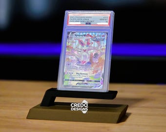 May include: A graded Pokémon card featuring Sylveon VMAX from the Evolving Skies set, displayed in a clear protective case. The card is on a black and gold display stand. The text on the card reads "Max Harmony".
