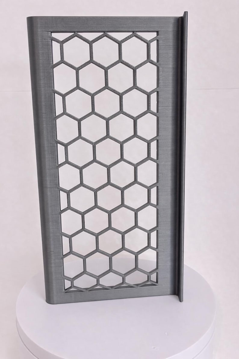 May include: A rectangular, dark gray panel featuring a repeating honeycomb lattice pattern. The structure has a solid frame with a vertical edge flange on the right side, displaying a textured surface finish consistent with 3D printed objects.