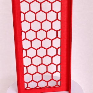 May include: A bright red rectangular panel featuring a repeating honeycomb pattern of open hexagonal cutouts. The structure has a solid frame border and a vertical side flange, displaying a textured surface finish consistent with 3D printing.