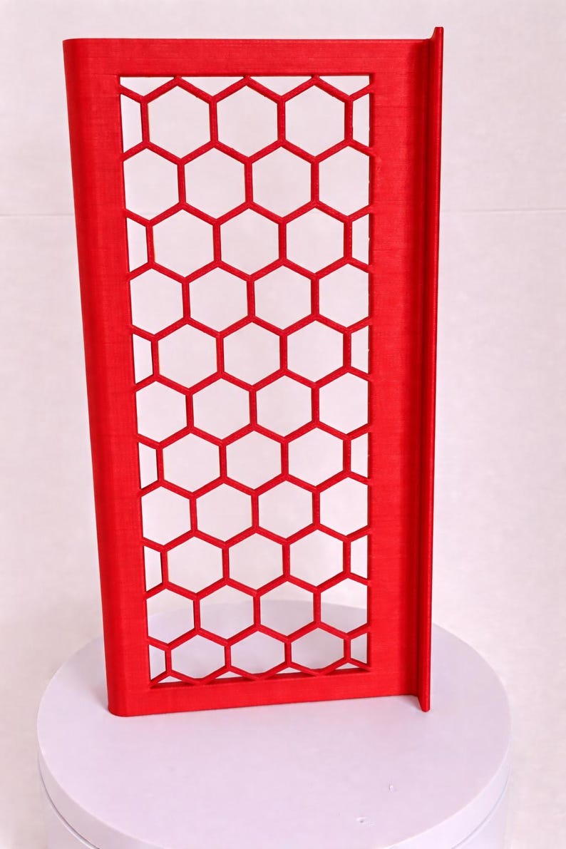 May include: A bright red rectangular panel featuring a repeating honeycomb pattern of open hexagonal cutouts. The structure has a solid frame border and a vertical side flange, displaying a textured surface finish consistent with 3D printing.