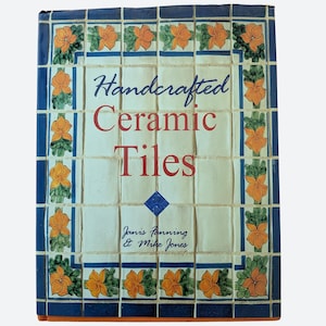 Op de afbeelding: Een hardcover boek getiteld Handcrafted Ceramic Tiles door Janis Fanning en Mike Jones. De kaft toont een raster van gebroken witte tegels met een blauwe rand en een herhalend patroon van oranje bloemen en groene bladeren. Tekst in blauw en rood.